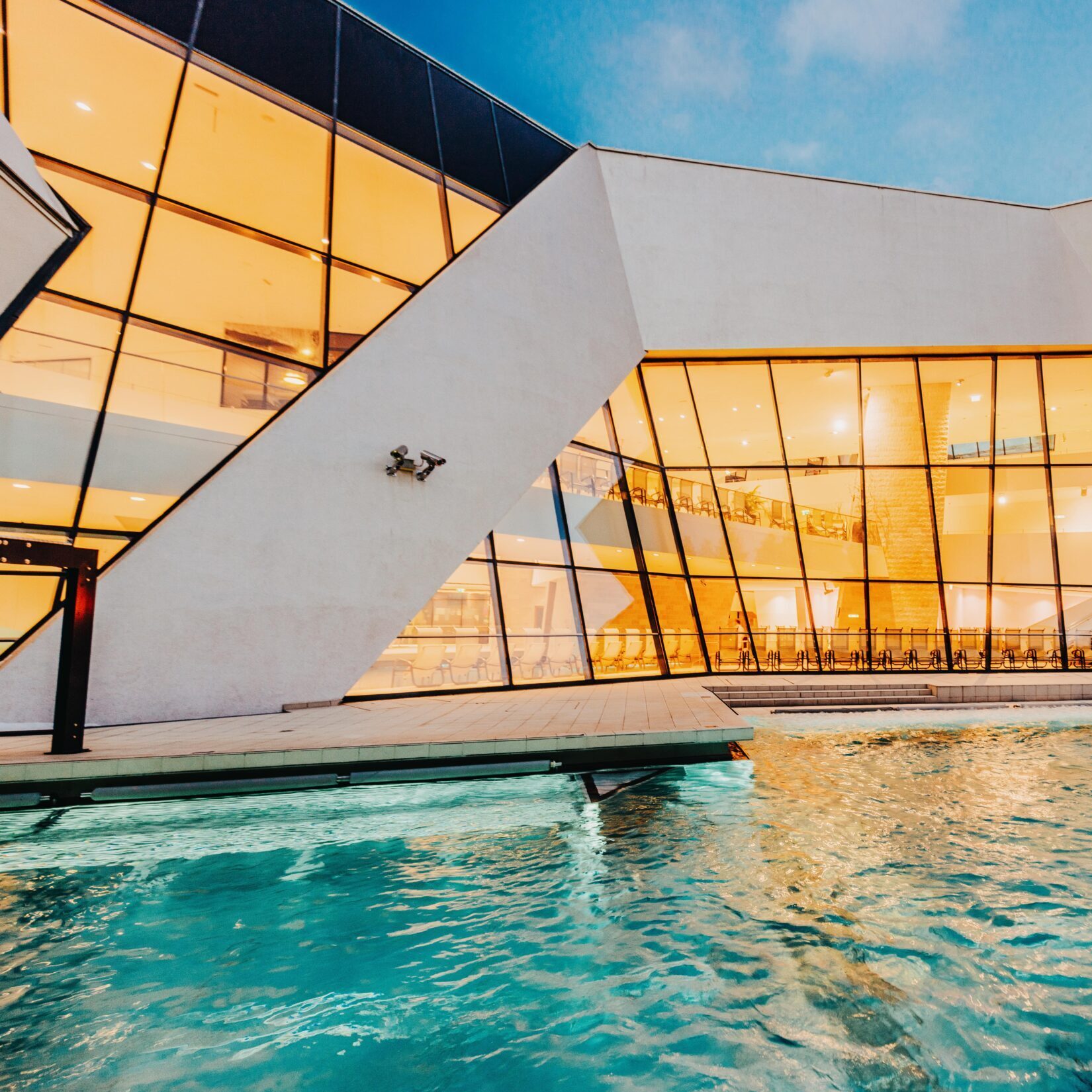Ein modernes Gebäude mit großen, eckigen Glasfenstern, die in der Abenddämmerung beleuchtet werden und sich in einem Außenpool mit klarem, blauem Wasser spiegeln - das Hotel Seestuben in Villach bietet eine atemberaubende Mischung aus modernem Design und ruhigem Seeambiente.