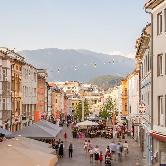 Eine belebte Fußgängerzone in Villach, gesäumt von bunten Häusern und Straßencafés; die Menschen flanieren und speisen in jedem der charmanten Restaurants, während sich im Hintergrund die Berge unter einem klaren Himmel erheben.